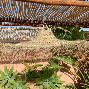 May include: A large, round, straw hat hangs from a wooden pergola covered in bamboo. The hat is made of natural fibers and has a long, flowing fringe. The hat is hanging over a lush green garden with a pink wall in the background.