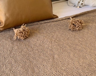 May include: A brown and black patterned throw blanket with two fluffy pom-poms. The blanket is on a bed with white and gray striped pillows and a brown leather pillow.