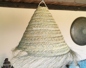 May include: A woven, conical lampshade made of natural fibers hangs from a wooden beam. The lampshade has a fringed bottom edge. The background includes a decorative metal plate, a lantern, and a seating area with blue cushions.