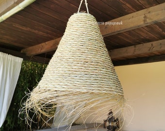 May include: A large, conical, woven lampshade hangs from the ceiling of a covered patio. The lampshade is made of natural fibers and has a light brown color. The patio is furnished with a bamboo bed and a small table.