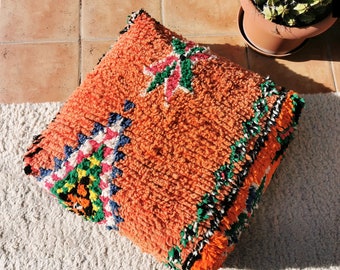 May include: A large, orange, fluffy pillow with a colorful geometric pattern. The pillow is sitting on a white, fluffy rug.