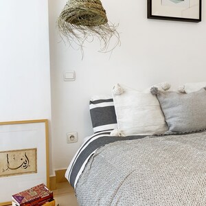 May include: A white bed with a gray and white patterned blanket and a woven straw lampshade hanging from the ceiling. The bed has a white headboard and a gray and white striped pillow. There is a framed print on the wall behind the bed.