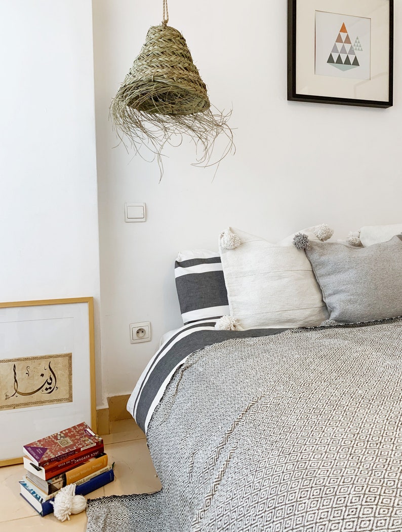 May include: A white bed with a gray and white patterned blanket and a woven straw lampshade hanging from the ceiling. The bed has a white headboard and a gray and white striped pillow. There is a framed print on the wall behind the bed.