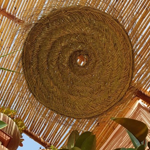 May include: A large, round, woven lampshade made of natural materials hangs from a bamboo ceiling. The lampshade is light brown and has a circular opening in the center. The bamboo ceiling is made of thin, light brown strips.