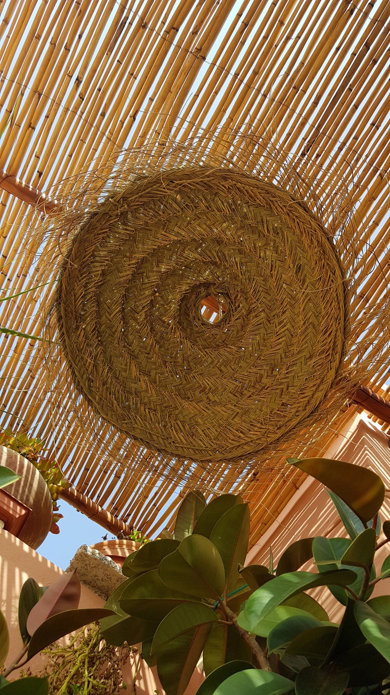 May include: A large, round, woven lampshade made of natural materials hangs from a bamboo ceiling. The lampshade is light brown and has a circular opening in the center. The bamboo ceiling is made of thin, light brown strips.