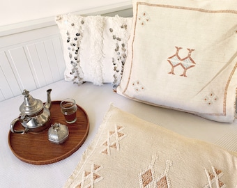 May include: A wooden tray with a silver teapot, a glass of tea, and a silver box with a decorative design. The tray is on a white bed with three pillows with embroidered designs.