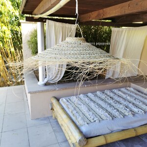 May include: A large, round, woven straw sun hat hangs from a wooden beam in a covered outdoor space. The hat is made of natural materials and has a wide brim. A white and gray striped blanket is on a bamboo bed in the background.