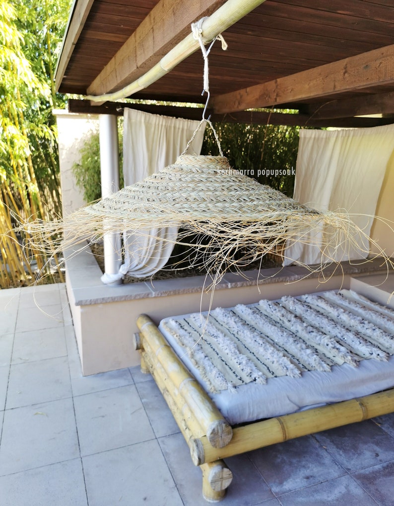 May include: A large, round, woven straw sun hat hangs from a wooden beam in a covered outdoor space. The hat is made of natural materials and has a wide brim. A white and gray striped blanket is on a bamboo bed in the background.