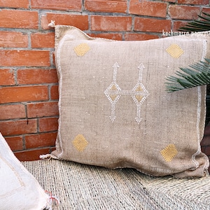 May include: A beige throw pillow with a geometric pattern in white and yellow. The pillow is made of a woven fabric and has a fringe border.