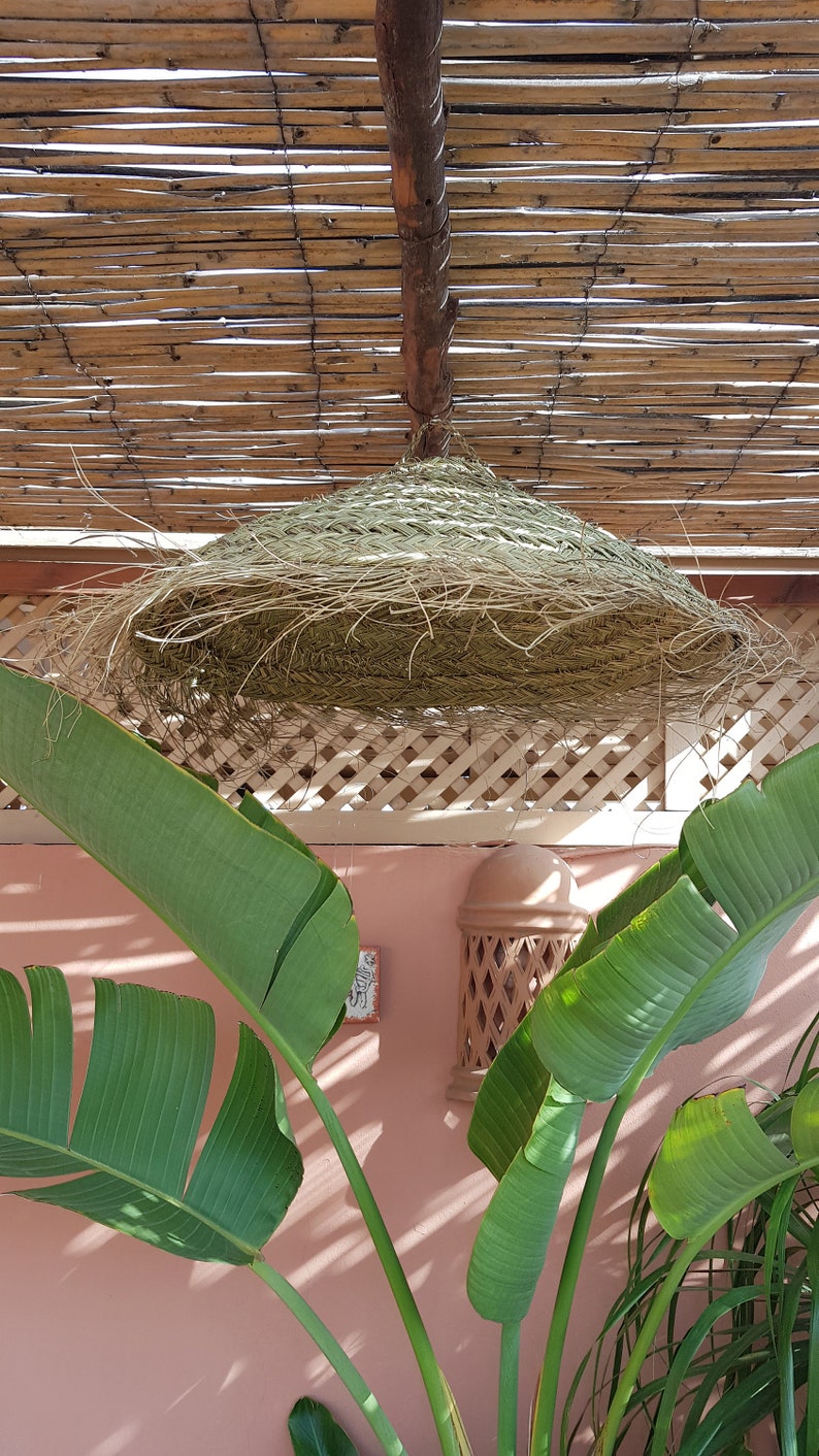 May include: A large, round, woven straw lampshade hangs from a bamboo ceiling. The lampshade is made of natural materials and has a rustic, bohemian style. The lampshade is hanging over a pink wall with green plants in the foreground.