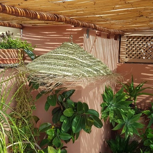 May include: A green woven sunshade hangs from a wooden pergola over a patio with potted plants. The patio is surrounded by a pink wall with a wooden lattice.