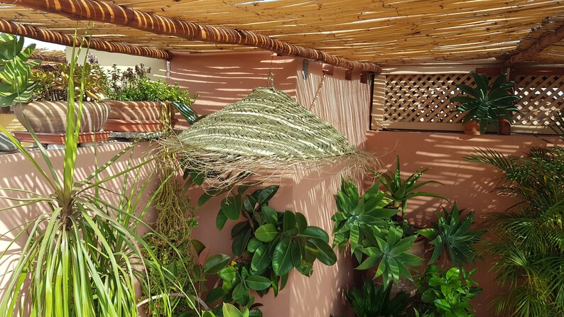 May include: A green woven sunshade hangs from a wooden pergola over a patio with potted plants. The patio is surrounded by a pink wall with a wooden lattice.