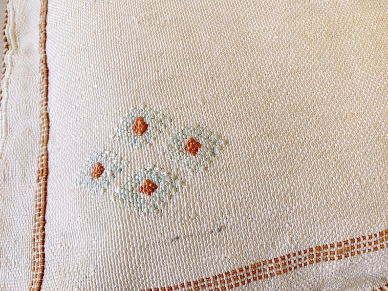 May include: A close-up of a white fabric with a brown border and a small embroidered design of three flowers in blue and orange.