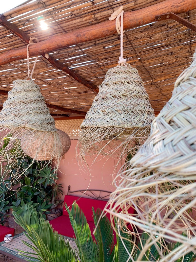May include: Three woven straw pendant lights hang from a bamboo ceiling. The lights are in a natural beige color and have a woven pattern. The lights are hanging over a red couch and a green plant.