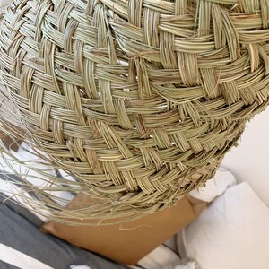 May include: A woven lampshade made of natural materials, with a light brown color and a textured surface. The lampshade is hanging over a bed with white and gray pillows.