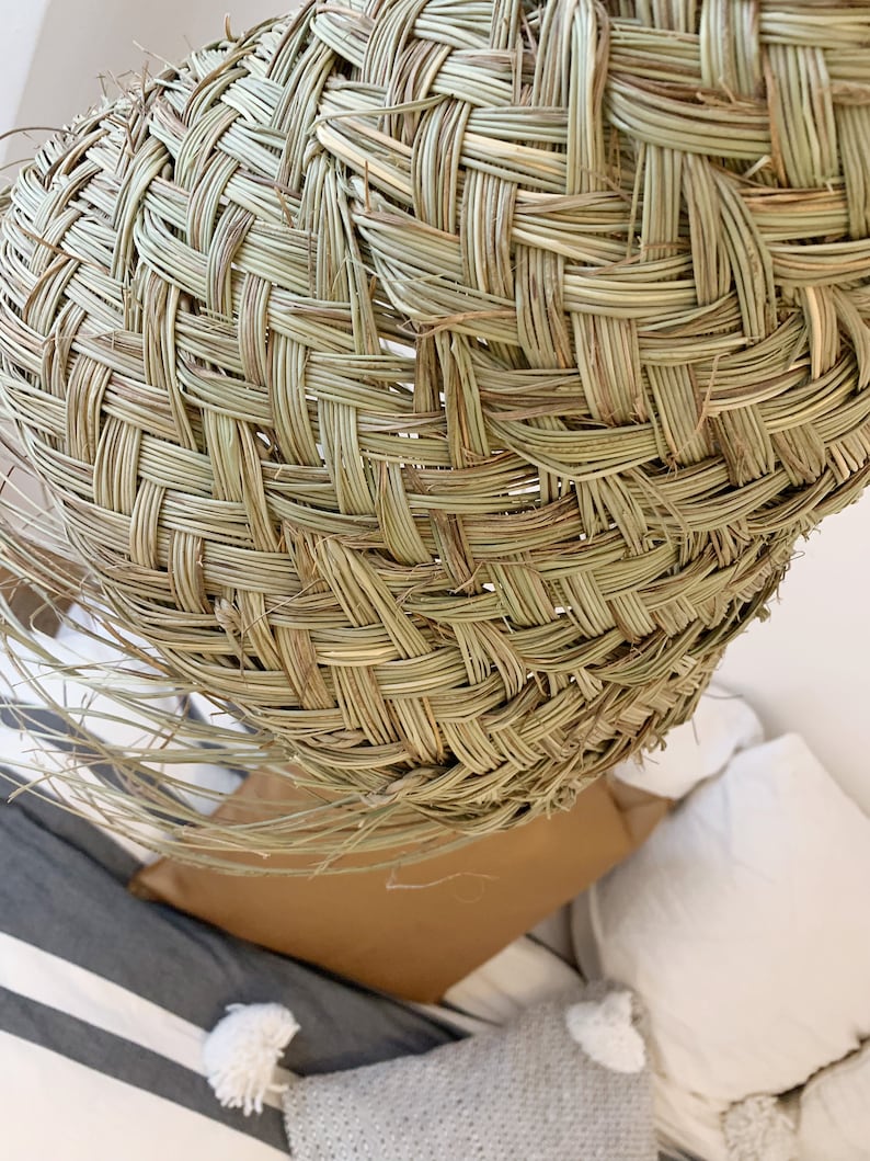 May include: A woven lampshade made of natural materials, with a light brown color and a textured surface. The lampshade is hanging over a bed with white and gray pillows.