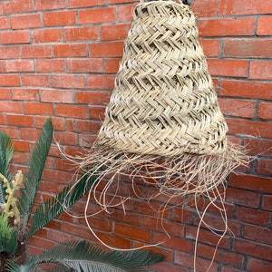 May include: A woven straw lampshade hanging from a metal hook against a brick wall. The lampshade is cone-shaped and has a frayed edge.