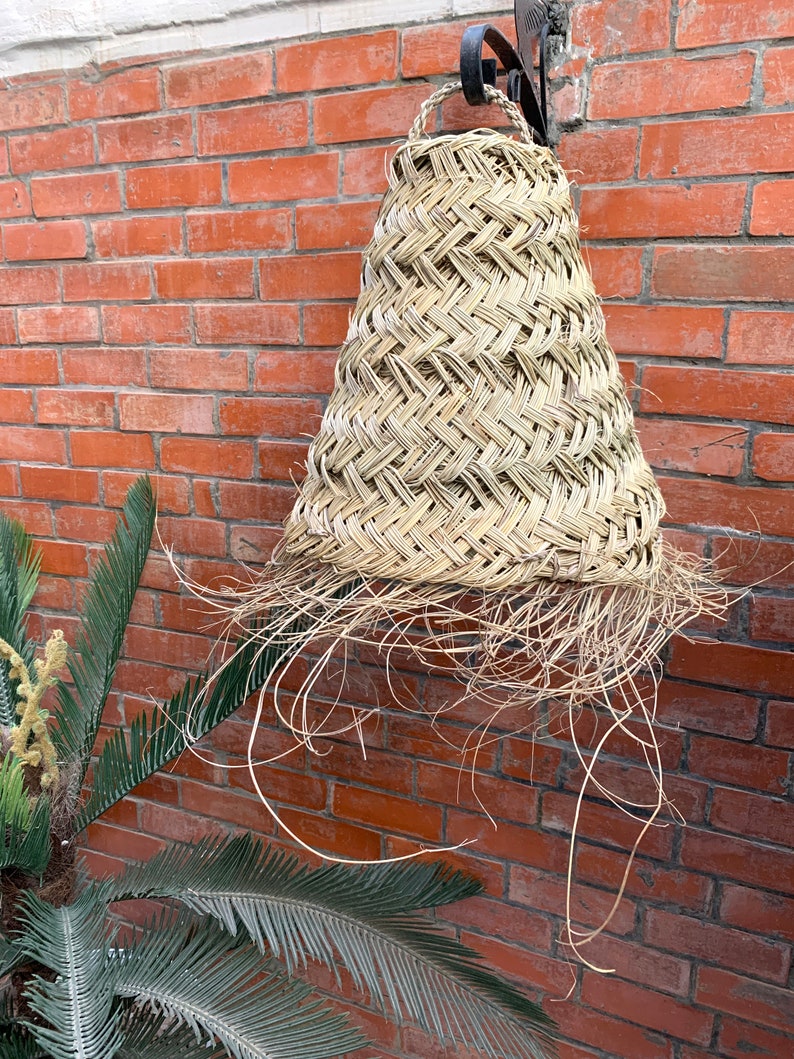 May include: A woven straw lampshade hanging from a metal hook against a brick wall. The lampshade is cone-shaped and has a frayed edge.