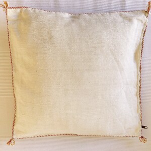 May include: A white pillow with a red trim and tassels. The pillow is made of a soft, woven fabric.