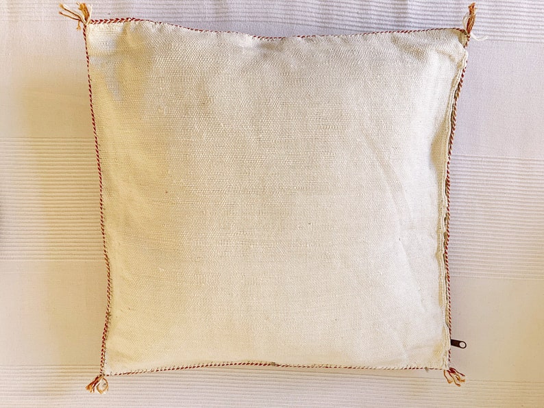May include: A white pillow with a red trim and tassels. The pillow is made of a soft, woven fabric.