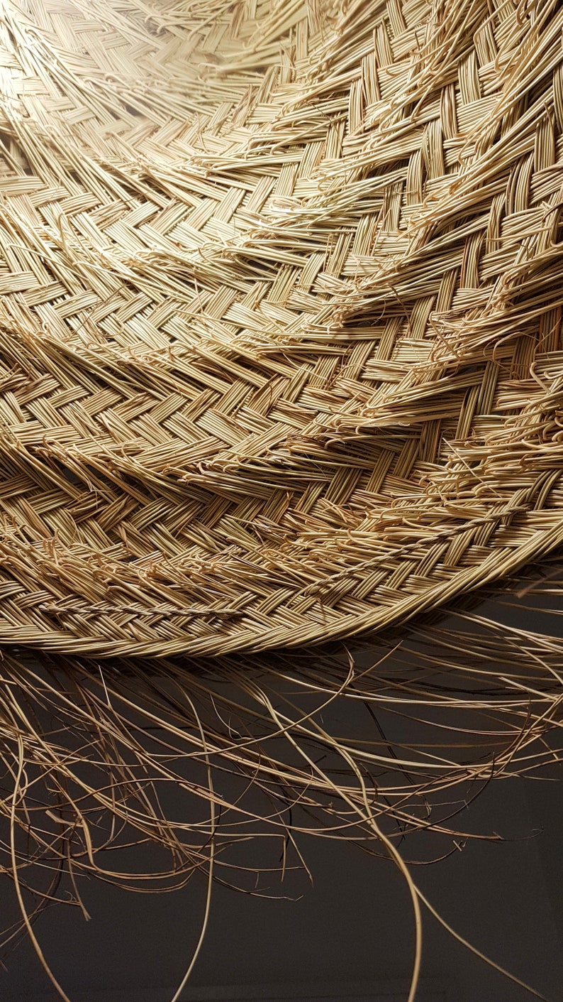 May include: Close-up of a woven straw hat with a natural brown color. The hat is made of tightly woven strands of straw, creating a textured surface. The hat is slightly curved, giving it a rounded shape.