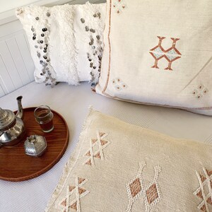May include: Three decorative throw pillows with embroidered geometric patterns in shades of beige and brown. The pillows are arranged on a white bedspread with a wooden tray and a silver teapot.