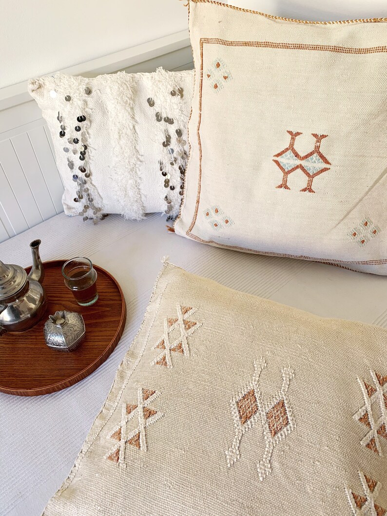 May include: Three decorative throw pillows with embroidered geometric patterns in shades of beige and brown. The pillows are arranged on a white bedspread with a wooden tray and a silver teapot.