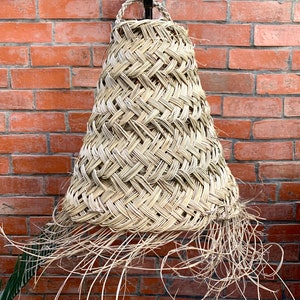 May include: A woven, natural fiber lampshade with a fringe detail hangs from a hook against a brick wall.