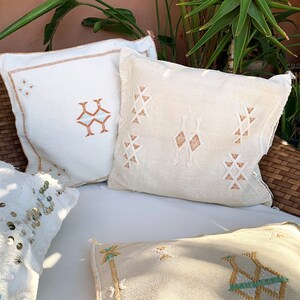 May include: Four decorative throw pillows with embroidered geometric patterns in shades of white, beige, and brown. The pillows are arranged on a wicker chair with a white cushion.