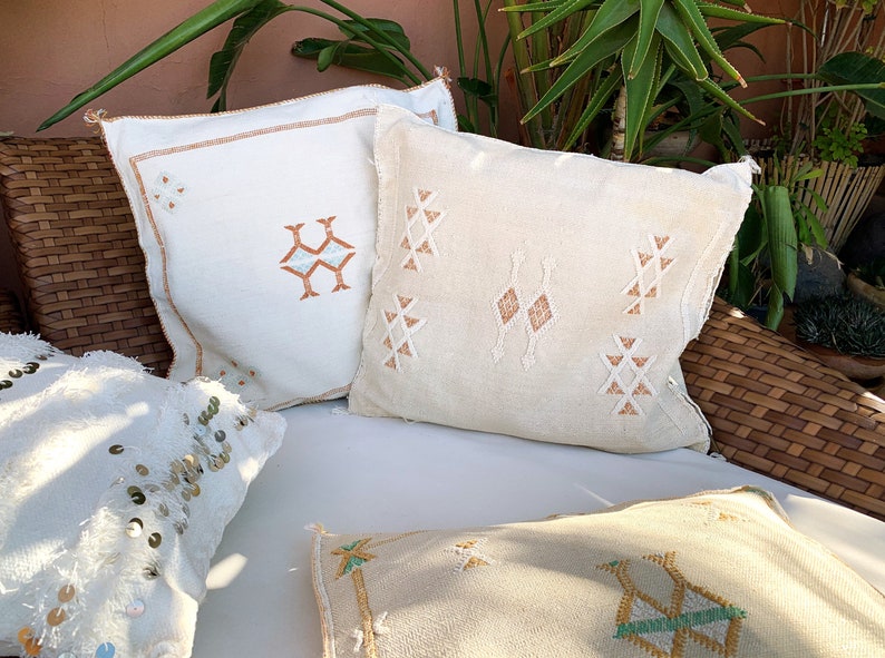 May include: Four decorative throw pillows with embroidered geometric patterns in shades of white, beige, and brown. The pillows are arranged on a wicker chair with a white cushion.