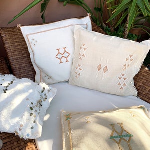 May include: Three decorative throw pillows with embroidered geometric patterns. The pillows are white with brown, beige, and green accents. The pillows are arranged on a wicker chair.