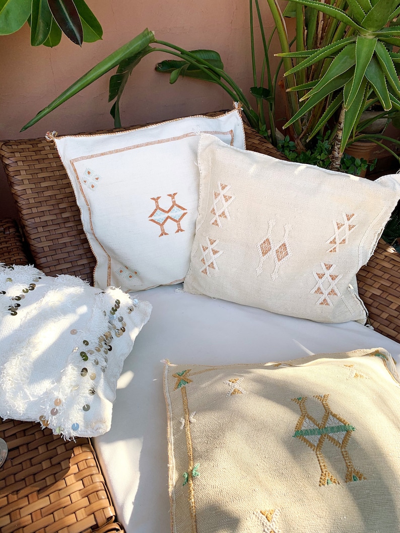 May include: Three decorative throw pillows with embroidered geometric patterns. The pillows are white with brown, beige, and green accents. The pillows are arranged on a wicker chair.