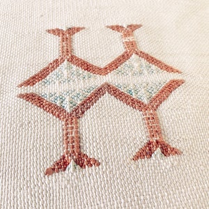 May include: Close-up of a white woven fabric with a geometric pattern in a light brown color. The pattern is made up of repeating shapes that resemble stylized birds.