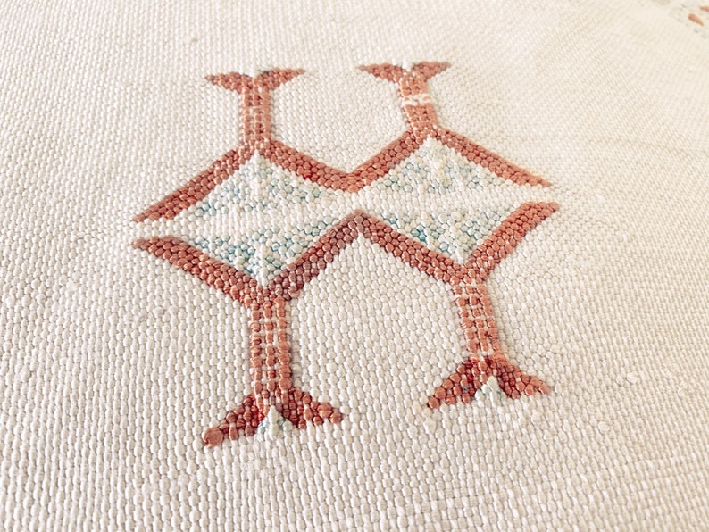 May include: Close-up of a white woven fabric with a geometric pattern in a light brown color. The pattern is made up of repeating shapes that resemble stylized birds.