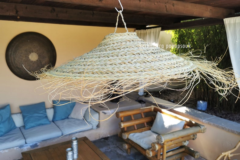 May include: A large, woven, straw lampshade hangs from the ceiling of a patio. The lampshade is light brown and has a wide brim. The patio is furnished with a couch and a chair.