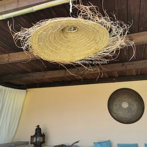 May include: A large, round, woven, straw lampshade hangs from a wooden ceiling. The lampshade is made of natural materials and has a rustic, bohemian style.