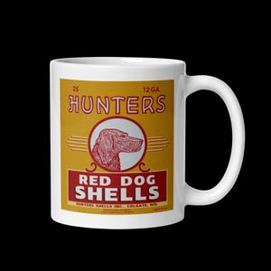 May include: A white ceramic mug featuring a vintage-style graphic of a dog in a red circle. The text reads 25, 12 GA., HUNTERS, RED DOG SHELLS, HUNTERS SHELLS INC., COLGATE, WIS., SMOKELESS POWDER, and NON-CORROSIVE PRIMER in red and yellow tones.