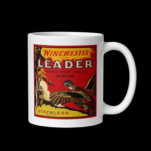 Könnte beinhalten: Ein weißer Keramikbecher mit einer Winchester Leader Vintage-Werbung. Die Grafik zeigt einen Jäger, einen fliegenden Vogel und den Text Winchester Leader Loaded Shot Shells Smokeless Staynless Made in U.S.A. auf rotem Hintergrund.