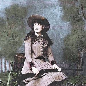 May include: A woman in a brown dress and a brown cowboy hat sits on a pile of hay, holding a rifle. She is wearing brown boots and has long dark hair. The background is a blurry forest.
