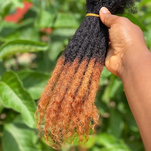 Puede incluir: Un paquete de rastas sintéticas con un esquema de color de dos tonos. La parte superior de las rastas es negra y la parte inferior es de color marrón claro. Las rastas son rizadas y parecen estar hechas de un material sintético suave.