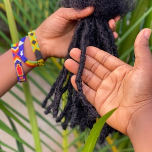 Peut inclure: Gros plan sur une main tenant un paquet de dreadlocks noirs. Les dreadlocks sont longues et épaisses, et elles sont disposées en un paquet propre et uniforme. La main porte plusieurs bracelets en perles colorées.