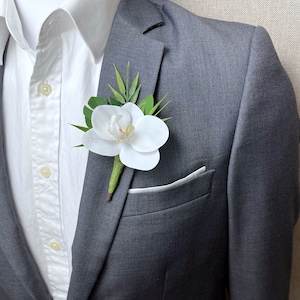 May include: A white orchid boutonniere with green leaves pinned to the lapel of a grey suit jacket worn over a white button-down shirt. The text Adult Design #2 is visible at the bottom of the image, featuring a floral arrangement on a suit.