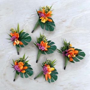 May include: Six tropical flower boutonnieres with orange and pink flowers, green leaves, and a monstera leaf.