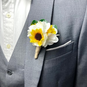 Puede incluir: Una chaqueta de traje gris con una camisa blanca abotonada debajo. Un pequeño ramo de flores blancas y amarillas está prendido en la solapa.