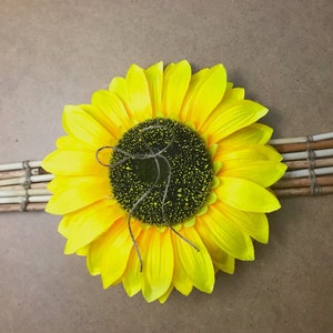 May include: A large artificial sunflower with bright yellow petals and a dark brown textured center, featuring a thin piece of twine tied in a bow on top. The flower is mounted horizontally across a small rectangular mat made of thin wooden sticks.