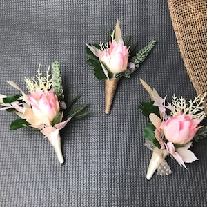May include: Three floral boutonnieres featuring pink flower buds, green foliage, and white accents. Each boutonniere is wrapped at the base with either tan twine or white ribbon, creating a structured arrangement of botanical elements for formal wear.