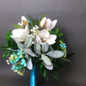 May include: A floral bouquet featuring white magnolia blossoms, fuzzy sage-green lamb's ear leaves, small white baby's breath, and bright turquoise filler flowers. The stems are tightly wrapped in a smooth, vibrant turquoise satin ribbon.