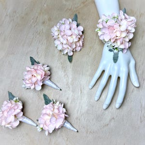 May include: Set of five pink hydrangea flower boutonnieres and wrist corsage. The boutonnieres are wrapped in white satin ribbon and the corsage is tied with a white satin ribbon.