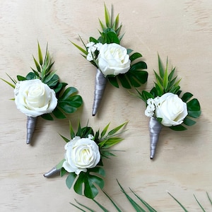 May include: Four floral boutonnieres, each featuring a single white rose, green monstera and grass-like foliage, and small white filler flowers. The stems are wrapped in a smooth, light gray satin ribbon, creating a uniform and structured appearance.