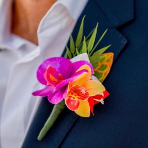 May include: A floral boutonniere featuring a vibrant magenta orchid and a yellow orchid with red speckled centers, accented by green palm-like leaves and a single orange-patterned leaf, pinned to the lapel of a dark navy blue suit jacket.